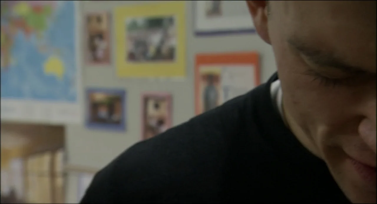 Hard as Nails (2001) movie still: closeup — A wall covered in various framed pictures and a world map hangs behind a man. The subject is a young man…