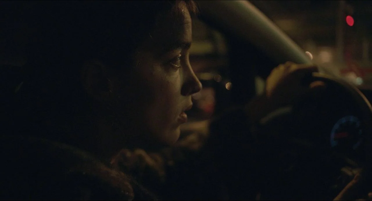 The Unknown Girl (2016) movie still: closeup — Inside a moving vehicle at night with blurred city lights visible through the window. A person with…