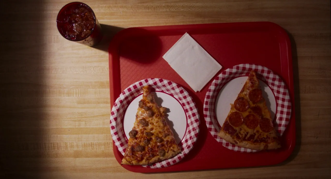 Drive-Away Dykes (2024) movie still: closeup — A top-down view shows a red plastic tray containing two slices of pizza and a napkin. The tray sits…