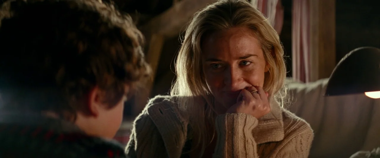 A Quiet Place (2018) movie still: medium — An attic space with dark wooden textures and soft ambient lighting from a lamp. A woman gazes at a young…