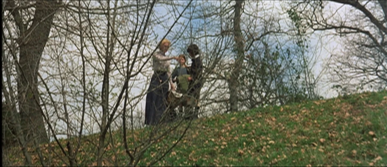 Flesh For Frankenstein (1973) movie still: fullbody — A grassy hill slope is populated by thin, leafless trees under a light sky. A woman and two…