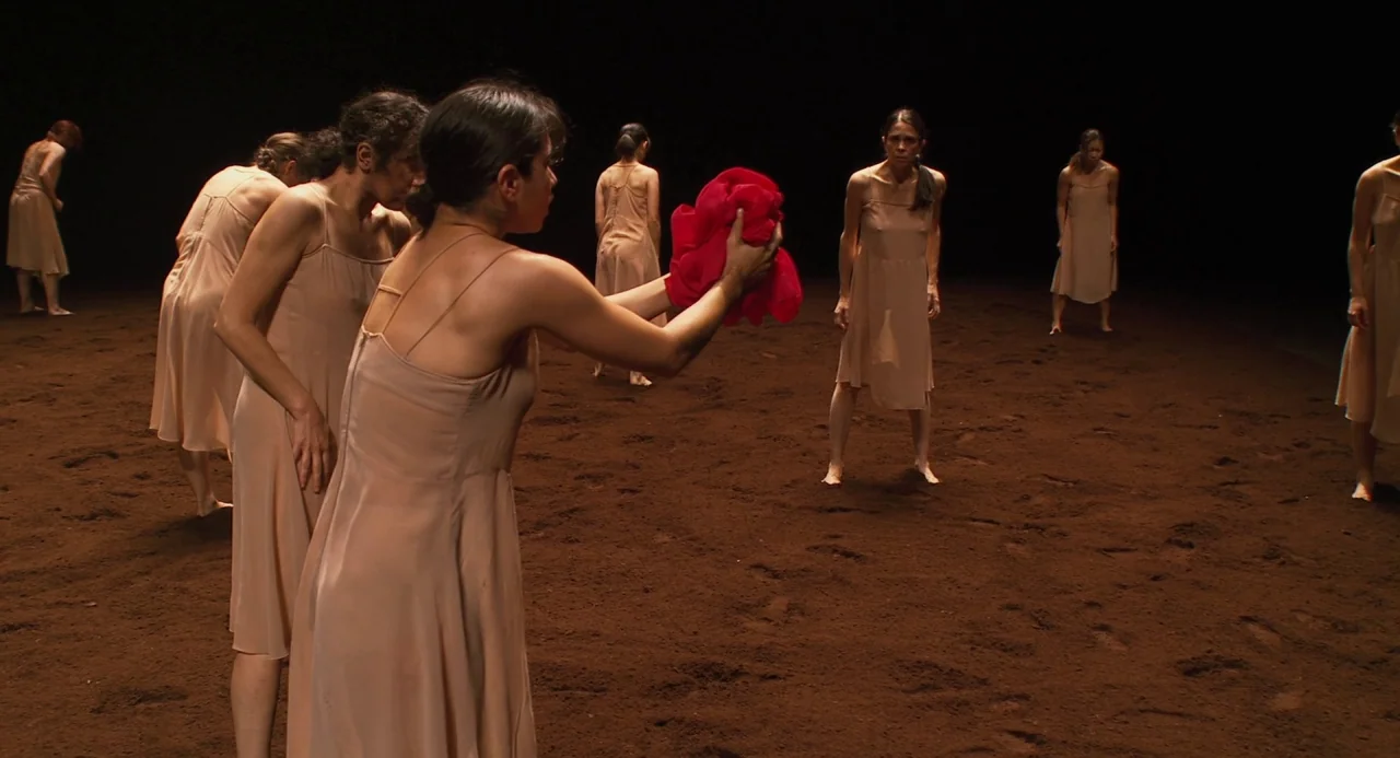 Pina (2011) movie still: wide — A group of women in light slip dresses stand on a dark, dirt-covered stage. One woman holds a bright red piece of…