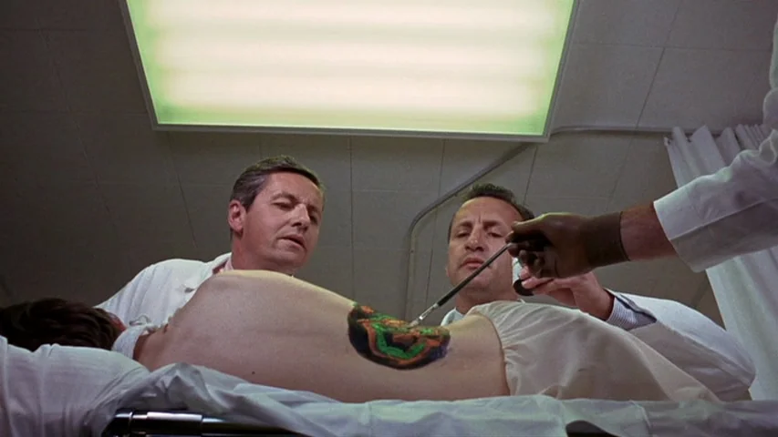 Petulia (1968) movie still: medium — A clinical examination room features a bright overhead fluorescent light panel and white privacy curtains. Two…