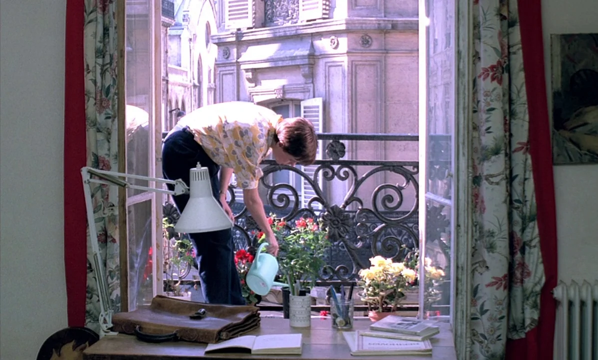 A Tale of Springtime (1990) movie still: medium — A light-filled room has a large window opening onto a wrought iron balcony. A woman in a patterned…