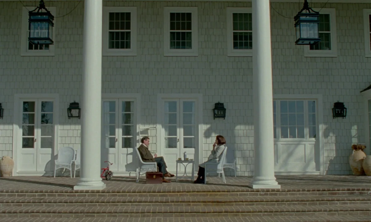 Jackie (2016) movie still: wide — A sprawling shingled estate house with white columns features a wide, paved front patio. Two people sit in white…