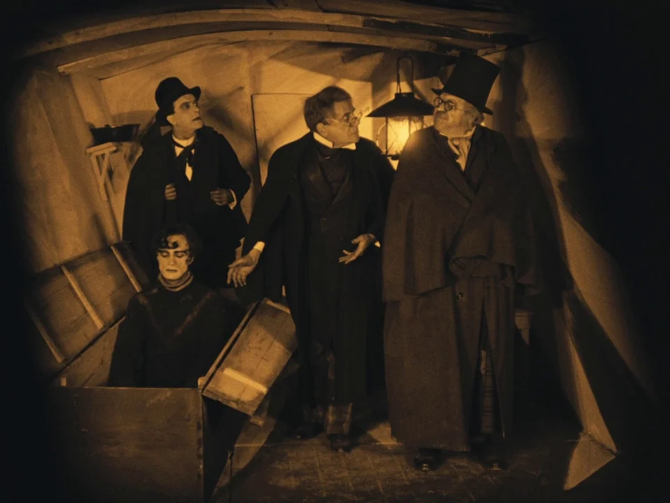 The Cabinet of Dr. Caligari (1920) movie still: fullbody — A dimly lit, cramped wooden cellar contains a coffin resting on the uneven floor. Four…