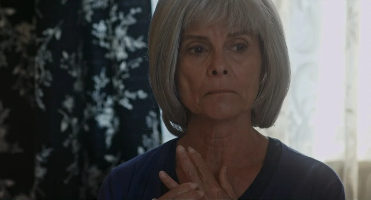 Gates of Darkness (2019) movie still: closeup — An interior space features a dark curtain with white floral patterns in the background. An elderly…