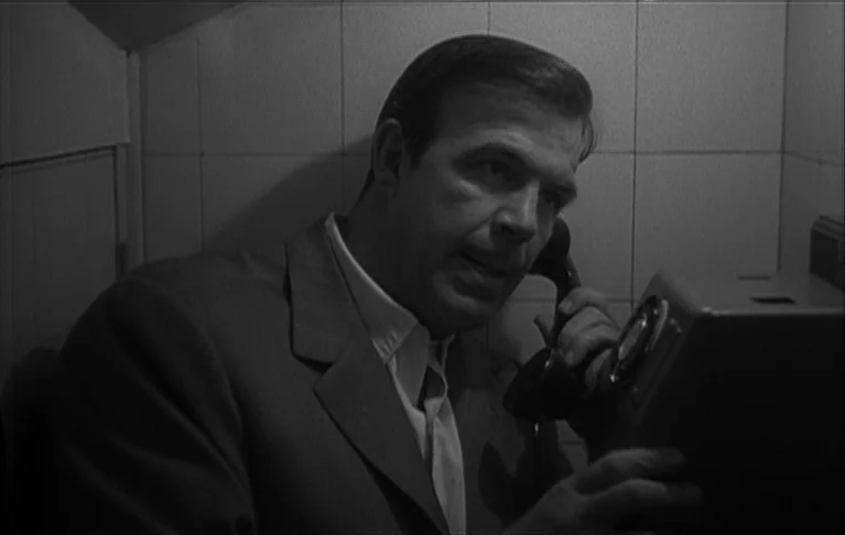 Le Signe du Lion (1962) movie still: medium — A man stands in a cramped, tiled corner while speaking on a wall-mounted phone. He holds the receiver…
