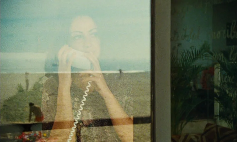 Documenteur (1981) movie still: medium — A woman is reflected in a pane of glass overlooking a sandy beach. She holds a white telephone to her ear…