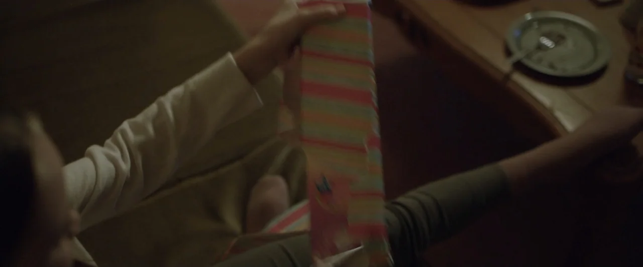 Motherly (2022) movie still: closeup — A dimly lit room contains a person holding a rectangular gift in their lap. A small plate with a fork rests…