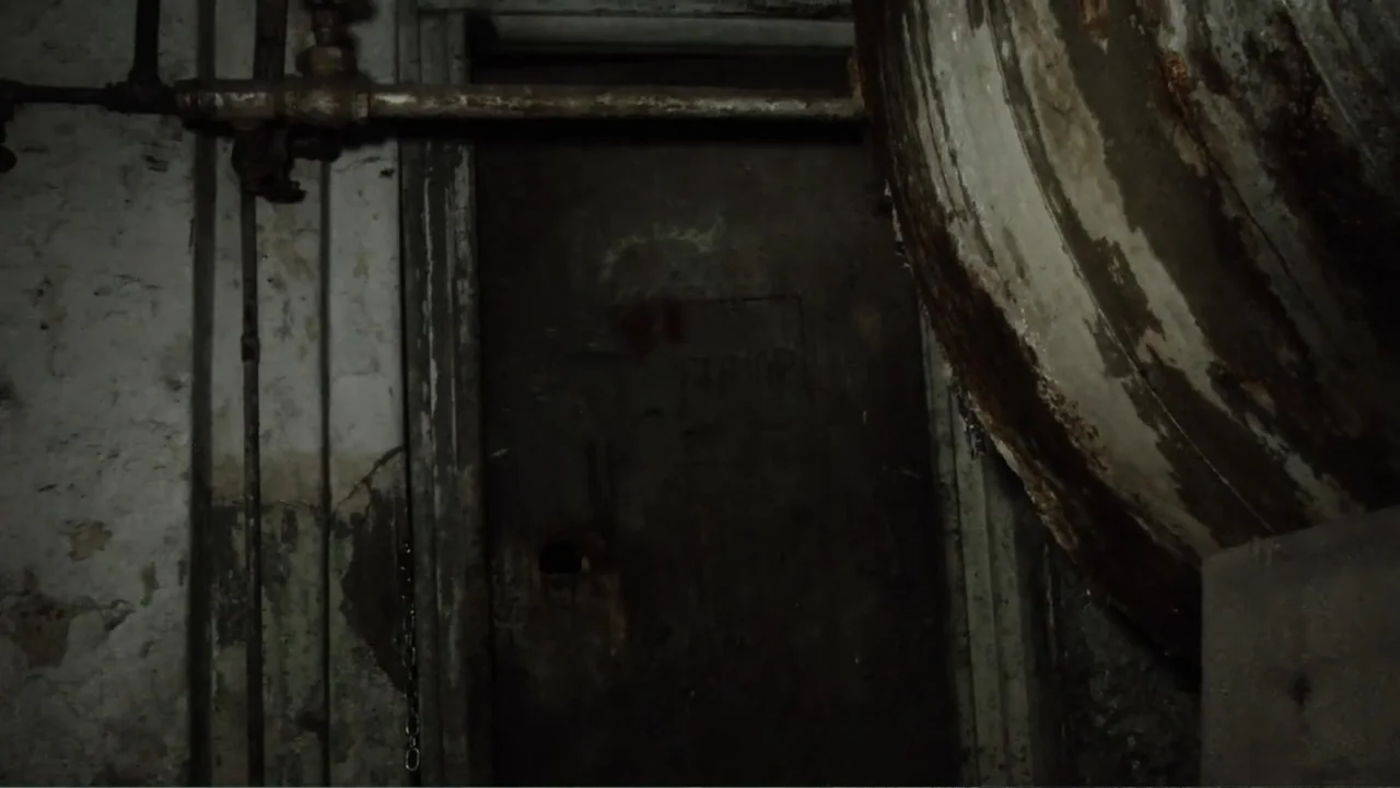 Lost (2010) movie still: medium — A dark industrial basement features a heavy metal door covered in rust and markings. A rusted pipe runs across the…