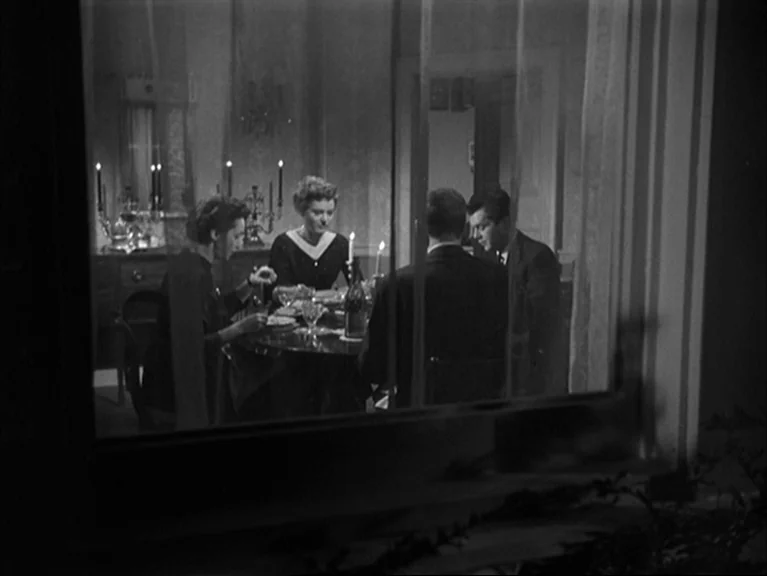 The Sleeping Tiger (1954) movie still: wide — A dimly lit room seen through a window shows four people dining at night. Two men and two women sit…