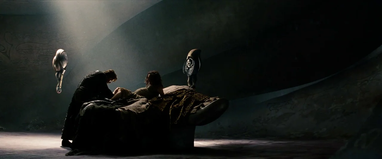 Man Of Steel (2013) movie still: wide — A dark, cavernous room with curved stone walls contains a low, monolithic stone bed platform. A man stands…