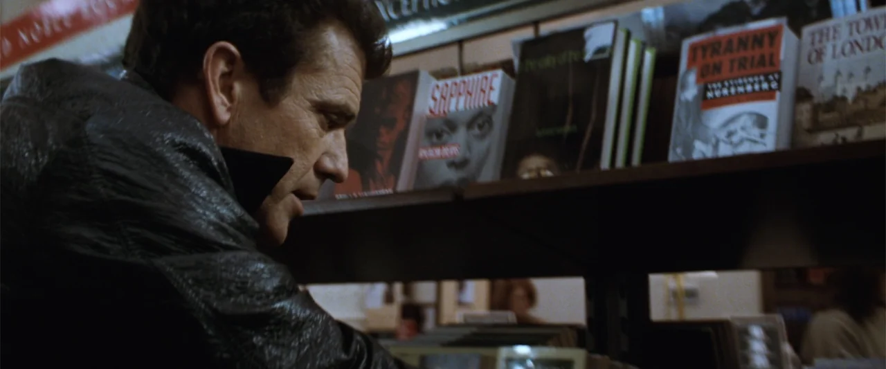 Conspiracy Theory (1997) movie still: medium — A man in a leather jacket stands before wooden shelves stocked with various books. He leans forward…