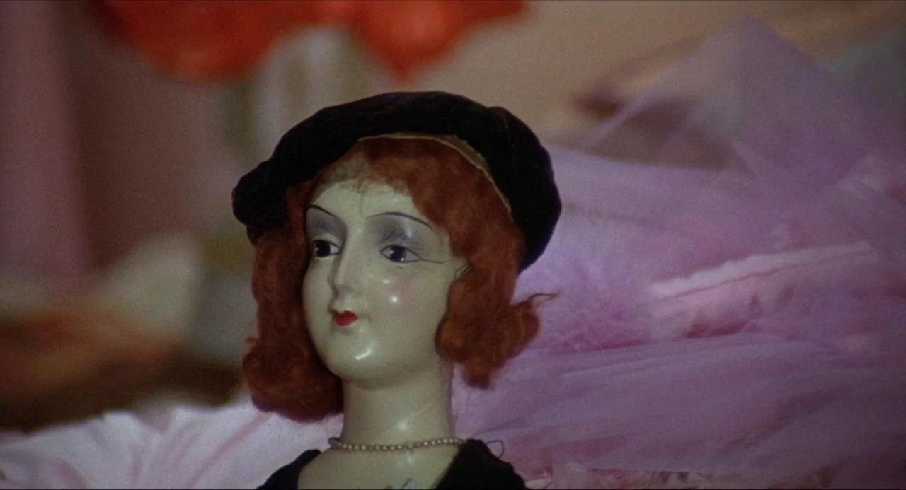 What’s the Matter with Helen? (1971) movie still: closeup — A close-up of a porcelain doll head positioned against layered pink tulle fabric. The…
