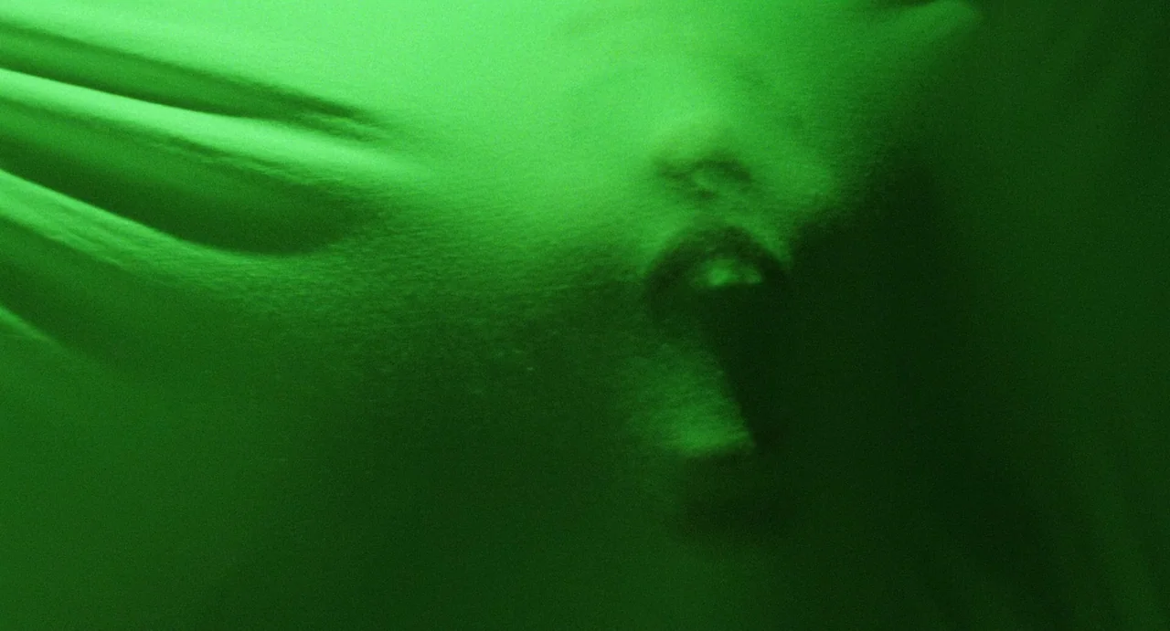 The Spider Labyrinth (1988) movie still: closeup — A person's face pushes against a thin, translucent green sheet from behind. The fabric stretches…
