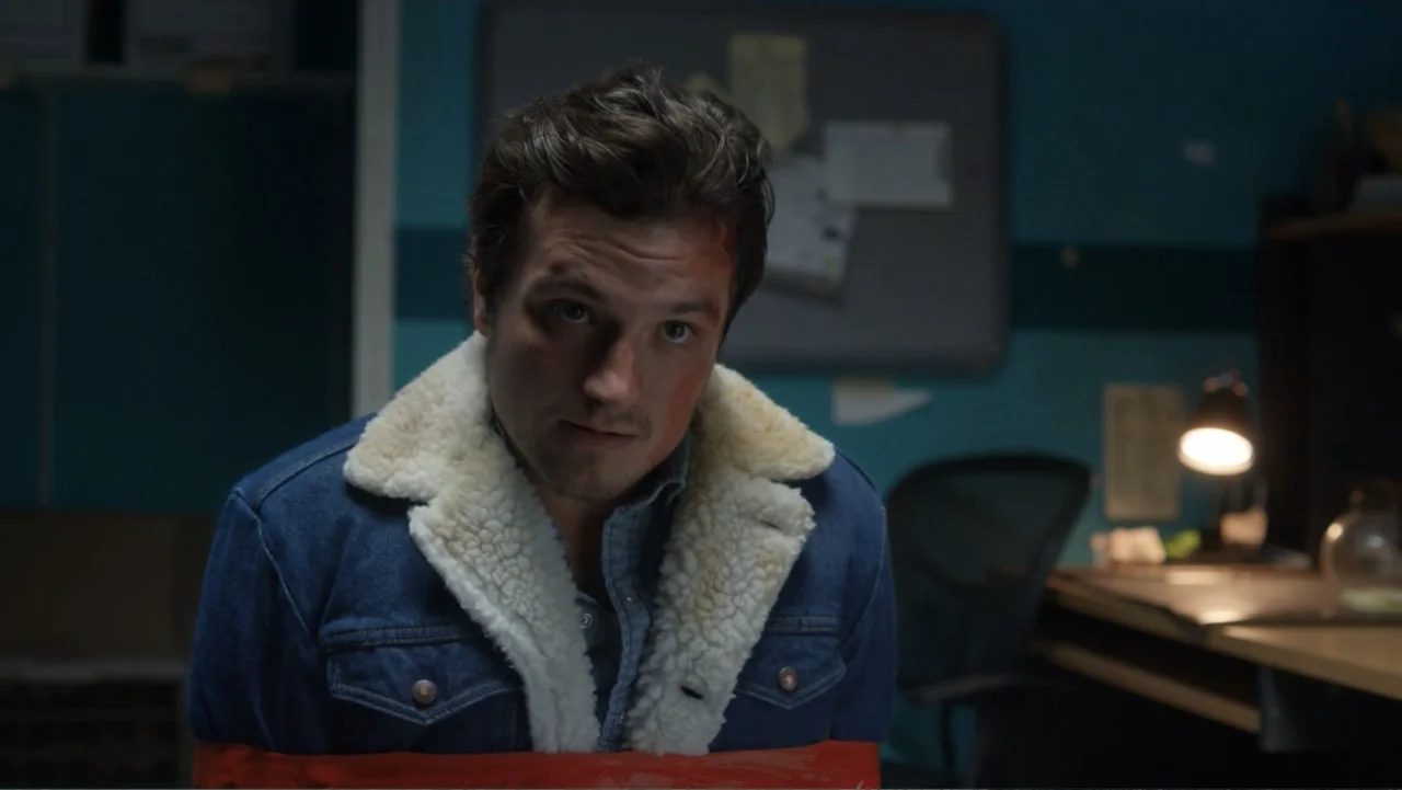 Burn (2019) movie still: medium — An office at night contains a desk with a glowing lamp and a bulletin board. A man with a bruised forehead sits…