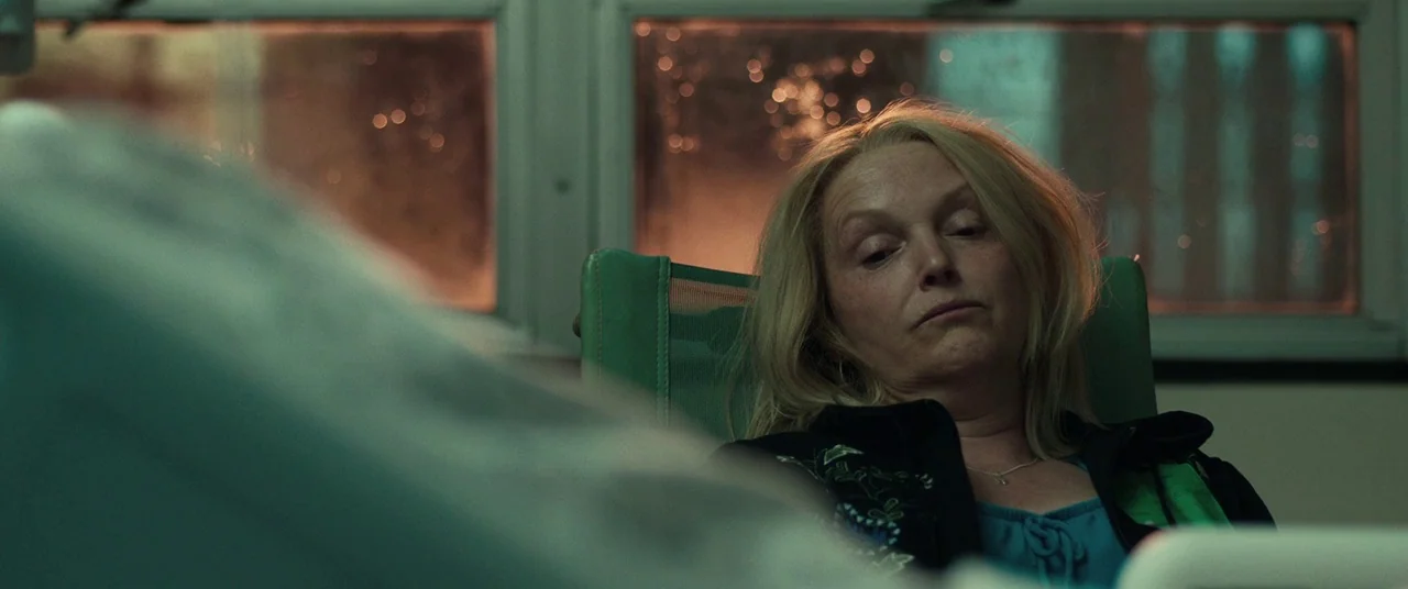 Stronger (2017) movie still: medium — A dimly lit room features a hospital bed and a green chair near a window. A woman sits in the chair and gazes…