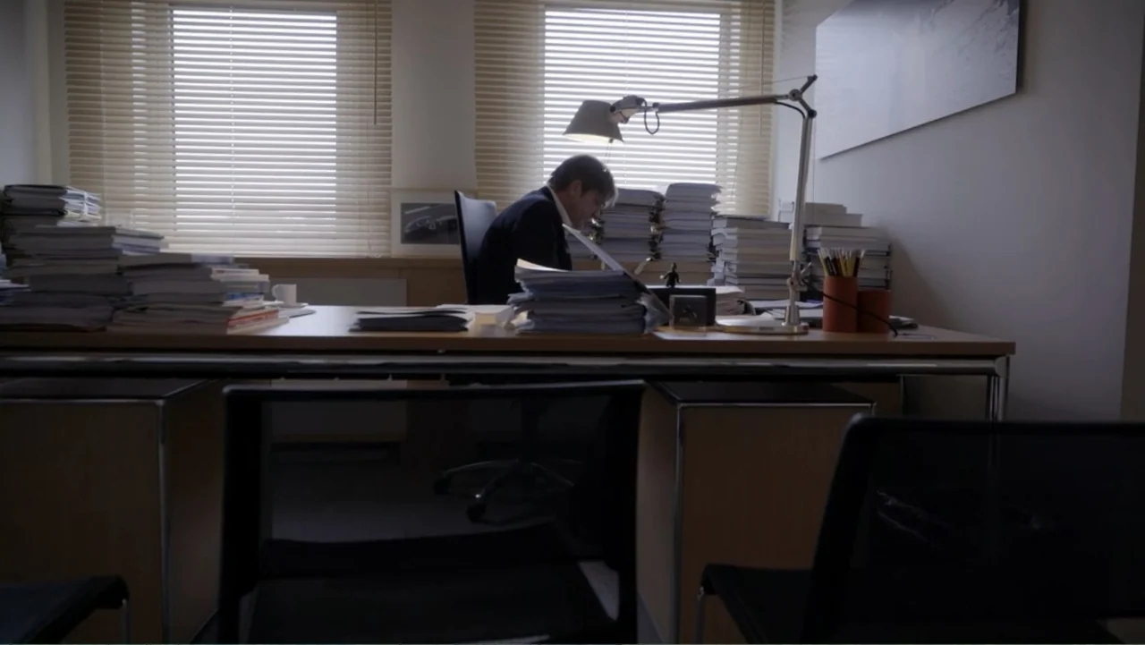Brexit: Behind Closed Doors (2019) movie still: wide — A cluttered, modern office space with multiple stacks of documents on the desk. A man in a…