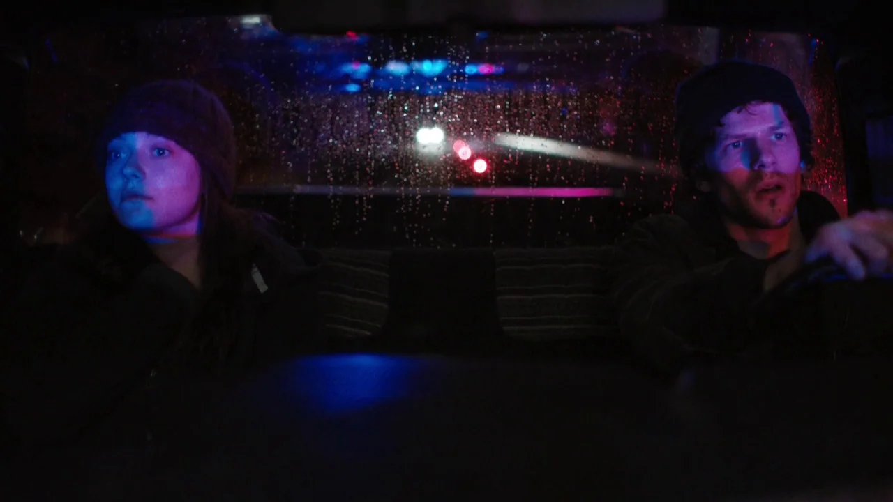 Night Moves (2013) movie still: medium — The interior of a moving vehicle at night with rain streaks on the windshield. A man and woman sit…