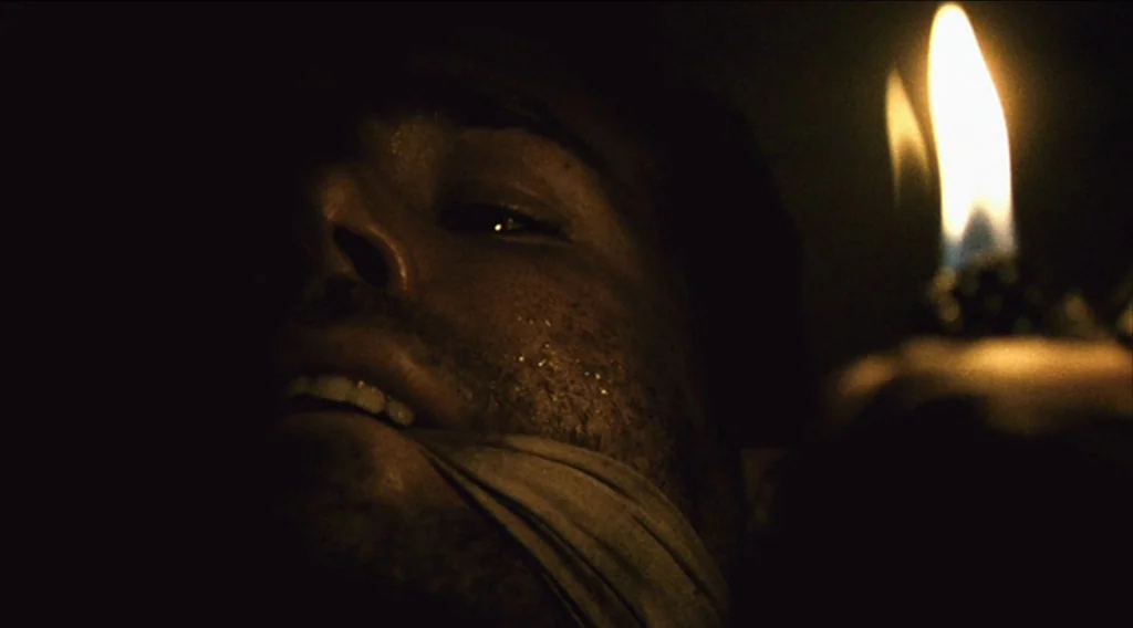 Buried (2010) movie still: closeup — A dark, enclosed space is illuminated only by a small, flickering flame. A man with stubble and a gag over his…