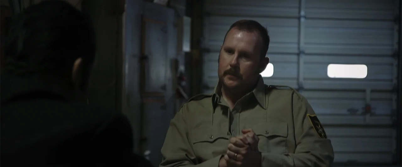 Darkness Waits (2020) movie still: medium — A dimly lit garage interior features a closed metal rolling door in the background. A man in a tan law…