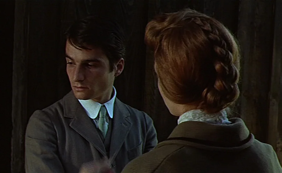 Two English Girls (1971) movie still: medium — Two individuals stand in a dim, wood-paneled room with shadows obscuring the background. The man in a…
