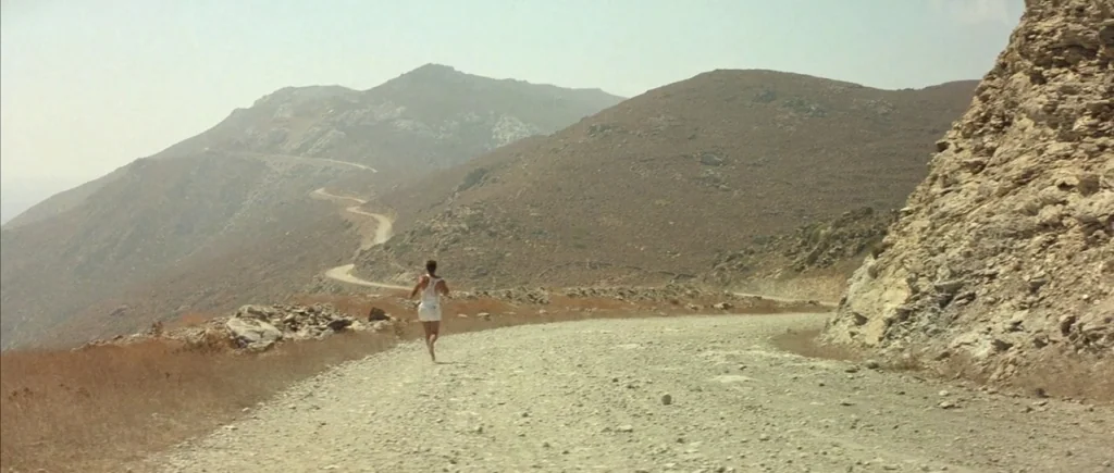 The Big Blue (1988) movie still: wide — A dry mountain range features a curving gravel road carved into the hillside. A lone runner travels down the…