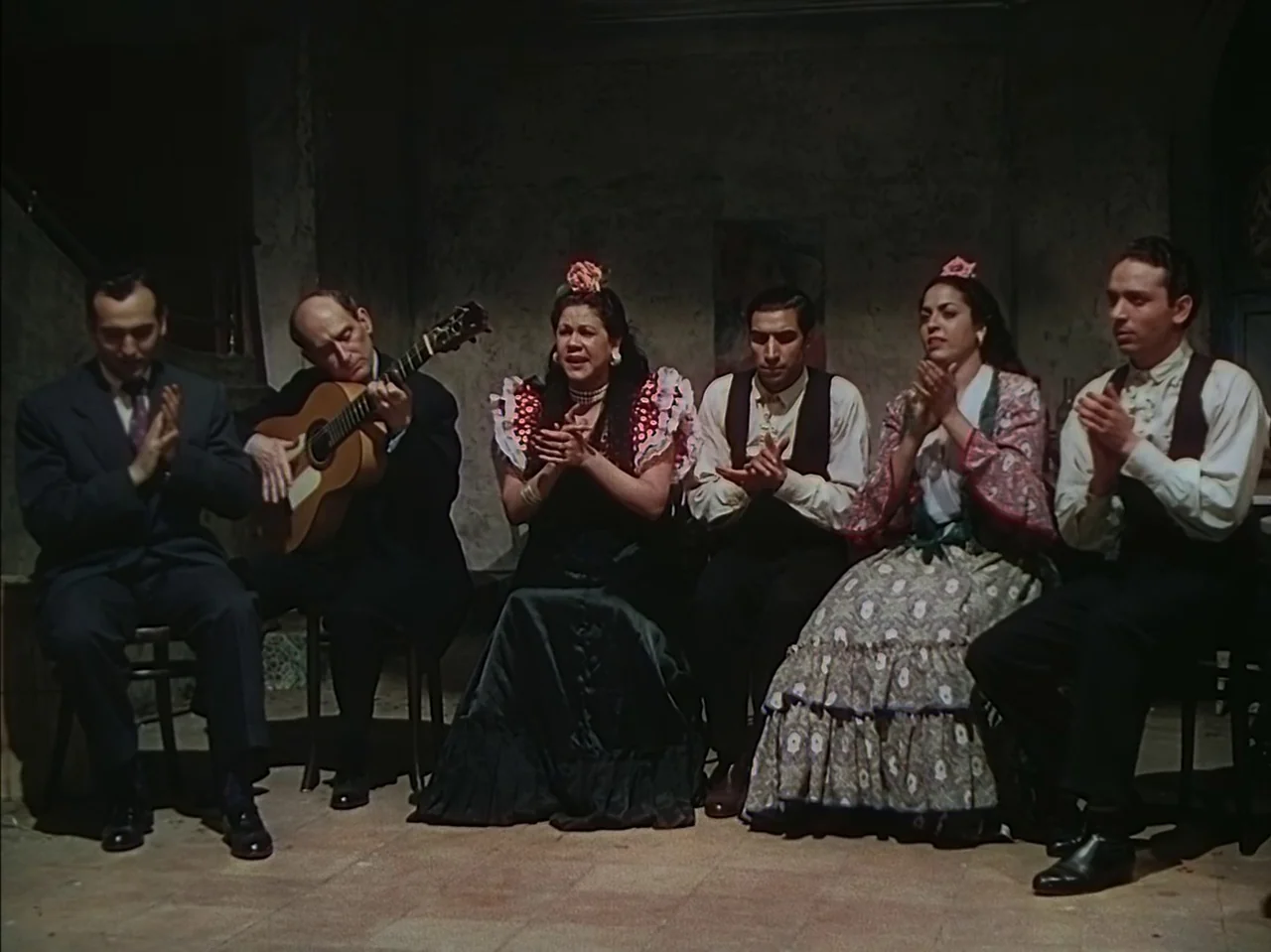 Pandora and the Flying Dutchman (1951) movie still: medium — A group of six people sits in chairs against a rough concrete wall. They are performing…