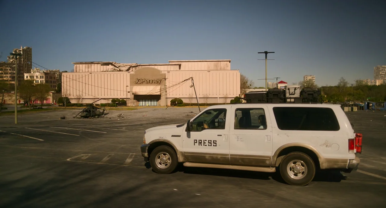 Civil War (2024) movie still: wide — An empty parking lot in front of a damaged JCPenney store building. A white SUV labeled press drives across the…