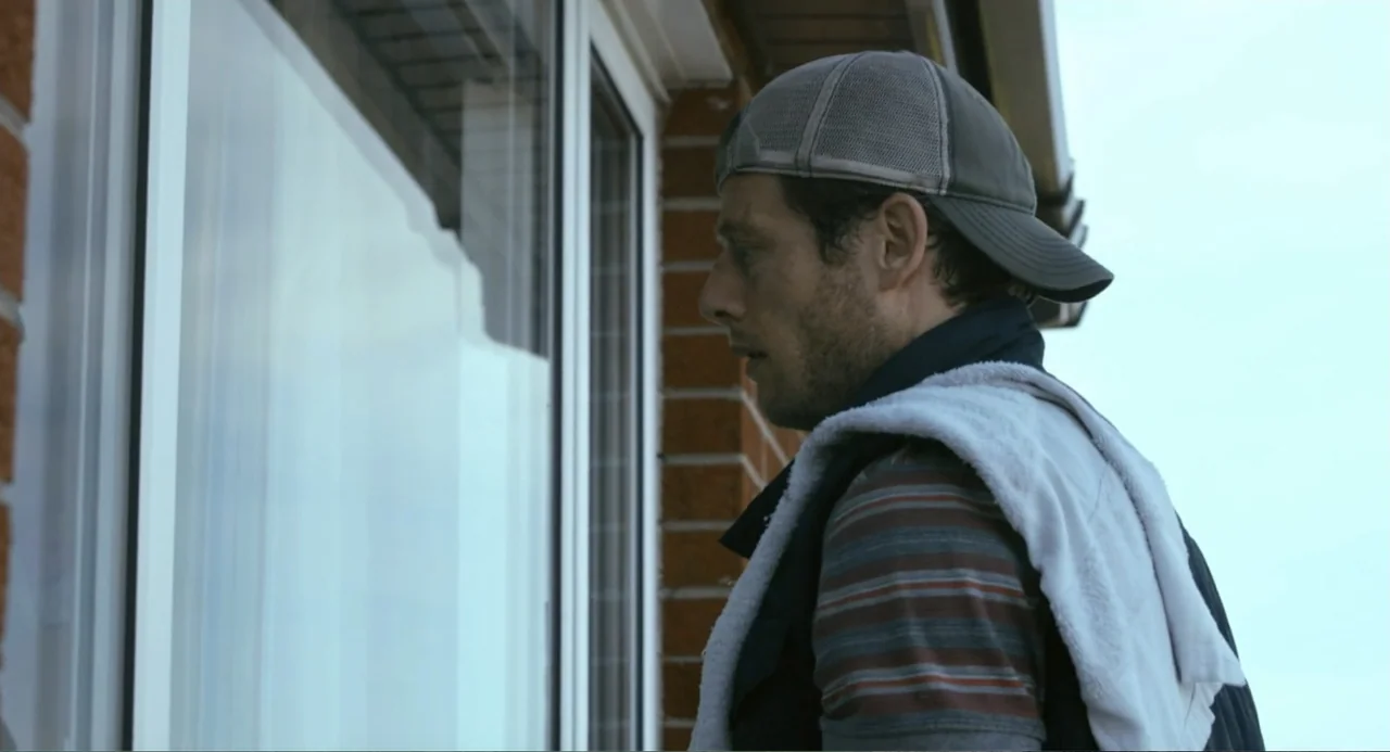 Living Out Loud (1998) movie still: medium — A man in casual clothing stands on a patio next to a glass window. He is facing the window, glancing at…