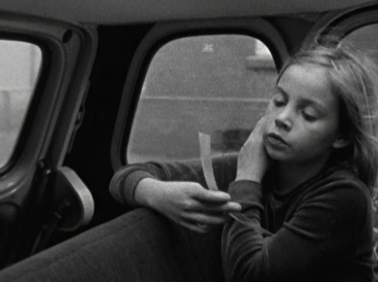 Alice in the Cities (1974) movie still: medium — The interior of a vehicle with a view of an out-of-focus street exterior. A young girl sits in the…