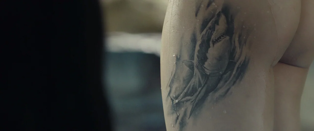 Beasts Clawing At Straws (2020) movie still: closeup — A close-up view shows a shark tattoo on wet skin in a shower setting. Droplets of water…