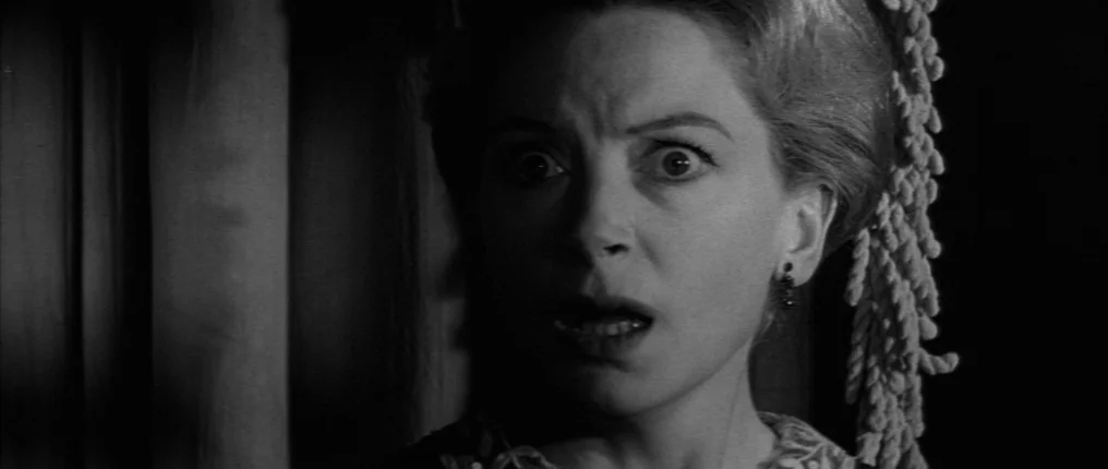 The Innocents (1961) movie still: closeup — A dimly lit indoor space with vertical drapery visible on the left side. A woman with light hair stares…