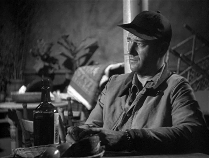 They Were Expendable (1945) movie still: medium — A dimly lit room contains a table with a bottle and other indistinct objects. A man in a cap sits…