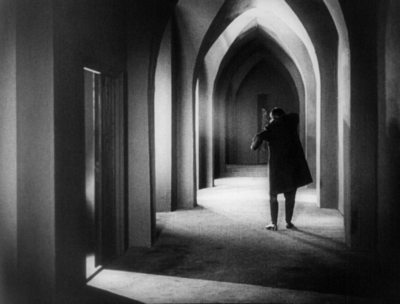 The Hands of Orlac (1924) movie still: wide — A long, cavernous stone hallway features repetitive pointed arches and deep, sharp shadows. A solitary…