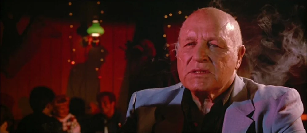 Reservoir Dogs (1992) movie still: closeup — A dimly lit club interior with red walls and blurred patrons in the background. An elderly bald man in…