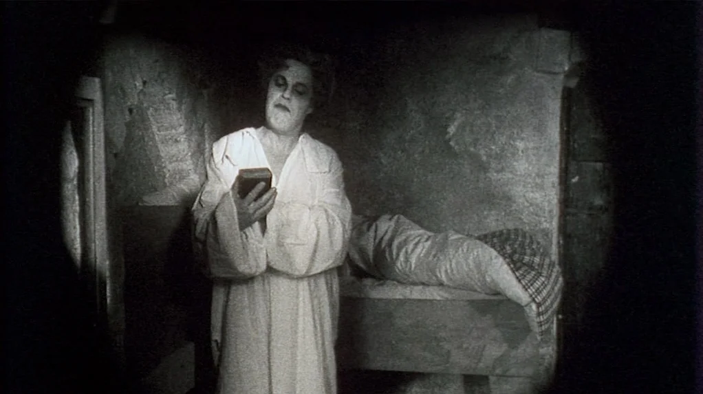 Shadow of the Vampire (2000) movie still: medium — A dark stone cell features a small raised cot with a rumpled blanket. A man in a nightgown holds…
