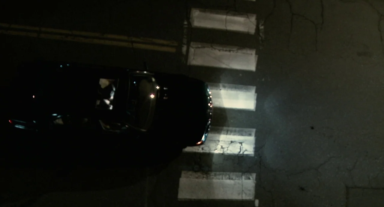 Another Earth (2011) movie still: wide — A top-down view of a dark asphalt road with a painted white crosswalk. A black sedan moves across the frame…