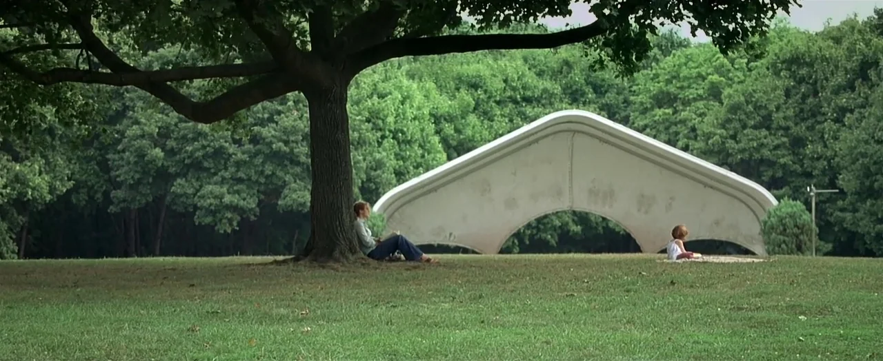Little Children (2006) movie still: wide — A green park with a large mature tree and a distinct arched white sculpture. An adult sits against the…