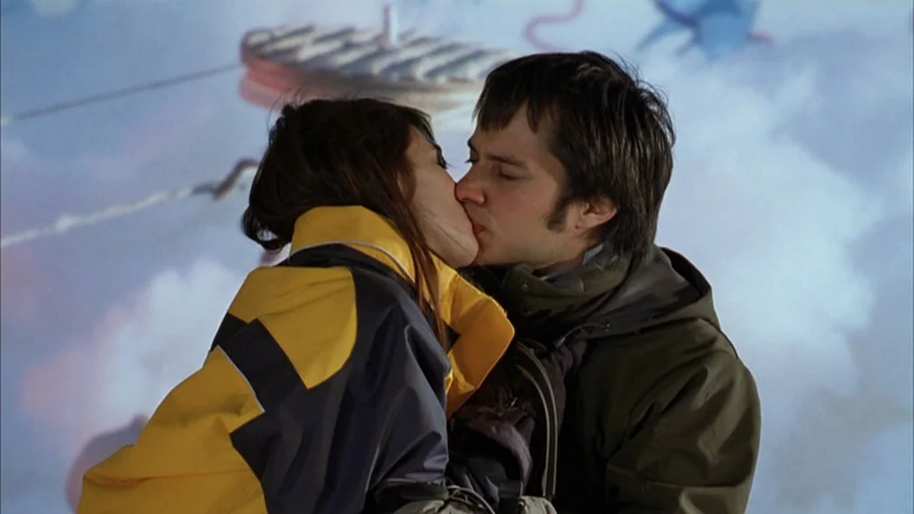 The Science Of Sleep (2006) movie still: medium — A man and woman kiss in front of a blue cloud projection screen. The subjects wear heavy outdoor…