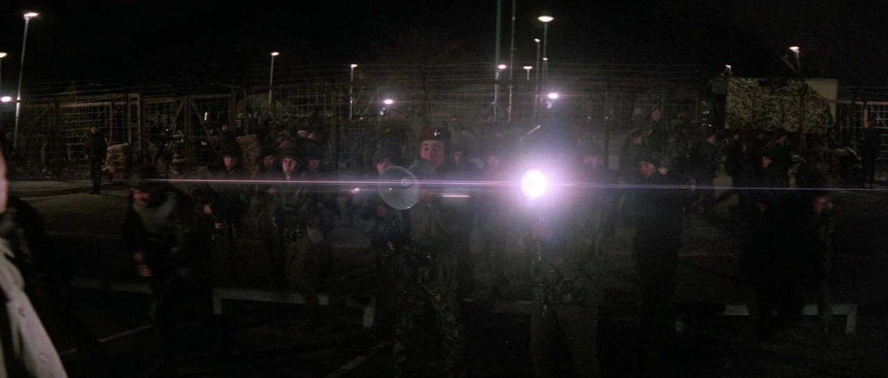 Lifeforce (1985) movie still: wide — A night-time military checkpoint with chain-link fencing, barbed wire, and bright floodlights. A soldier uses a…