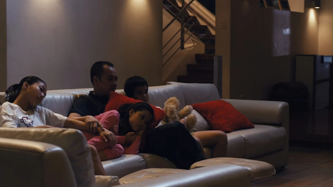Zero Tolerance (2015) movie still: medium — A modern interior living room features a white leather couch and a staircase background. A family of…