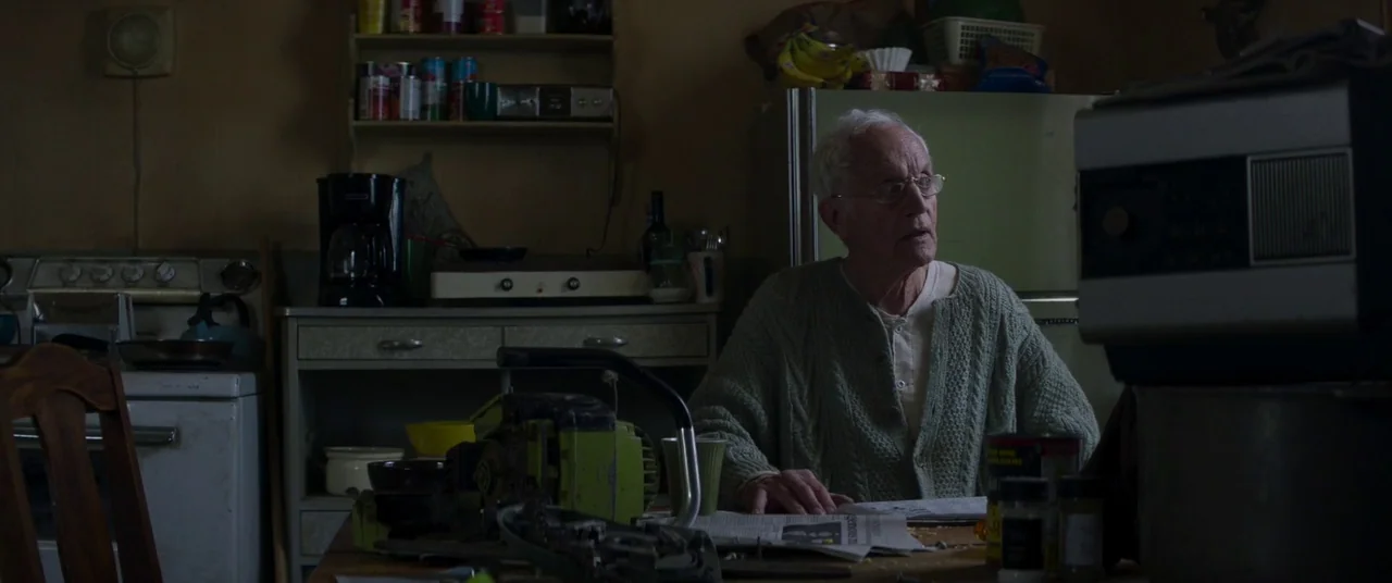 Falling (2020) movie still: medium — A dimly lit room with a cluttered table in the foreground and appliance-filled cabinetry. An elderly man sits…