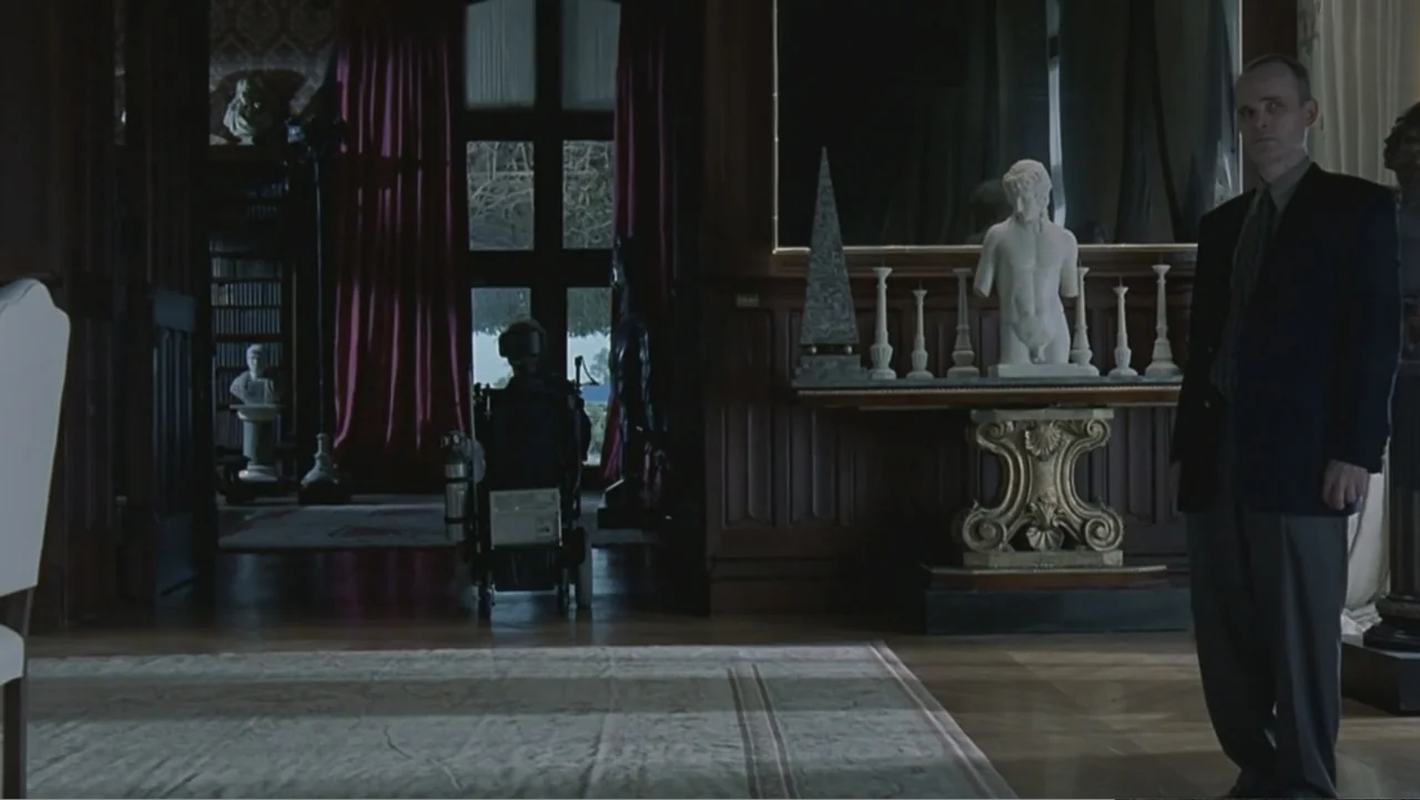 Hannibal (2001) movie still: medium — A wood-paneled library features a large mirror, classical marble busts, and a rug. A man in a suit stands on…