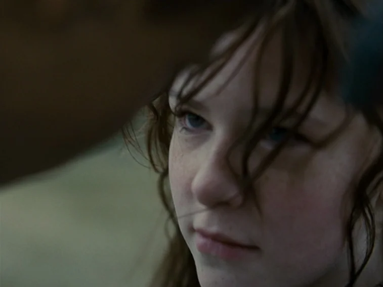 Wuthering Heights movie still: closeup — A close-up shot of a young girl with loose brown hair in a natural environment. The girl looks steadily at…