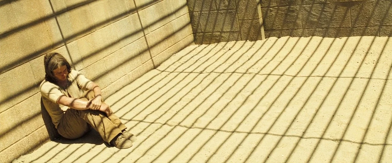 The Proposition (2005) movie still: wide — A corner of a stone enclosure features high walls and a sandy surface. A young man sits tucked into the…