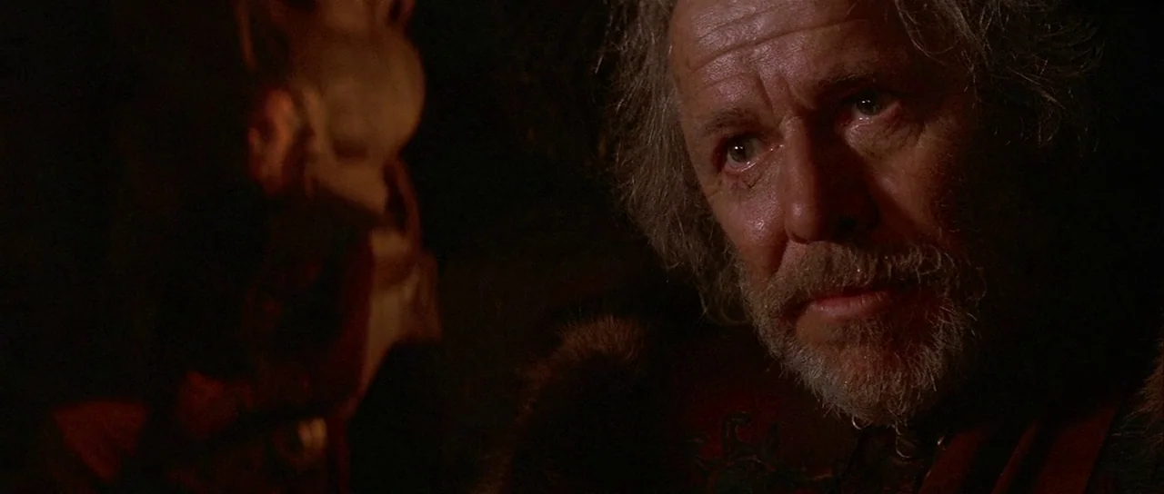 The 13th Warrior (1999) movie still: closeup — A dark, dimly lit cave interior provides a backdrop for the elderly man. He looks toward the side…