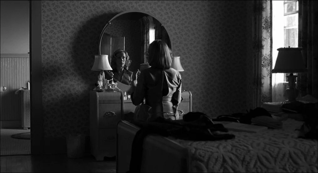 The Man Who Wasn’t There (2001) movie still: medium — A dark bedroom with patterned wallpaper, a round vanity mirror, and an unmade bed. A woman…