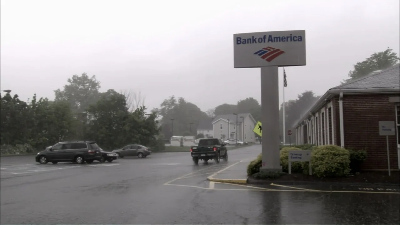 The Cheshire Murders (2013) movie still: wide — A wet bank parking lot under a gray, overcast sky during rainfall. Multiple cars occupy the paved…